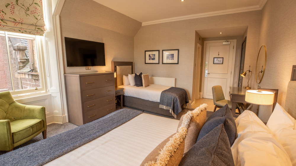 Room for 3 with a king-sized bed, a single bed on the opposite wall, a chair by a window, a dresser, a TV, and a door.