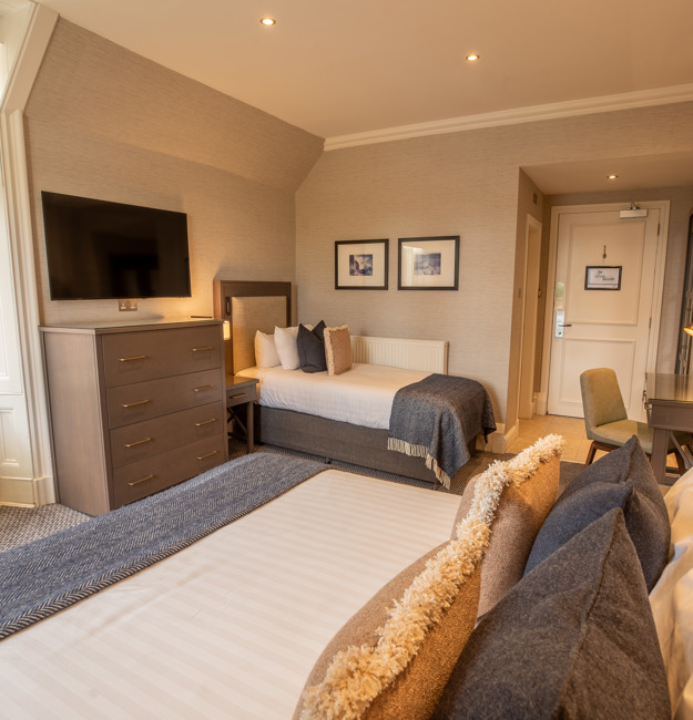 Room for 3 with a king-sized bed, a single bed on the opposite wall, a chair by a window, a dresser, a TV, and a door.