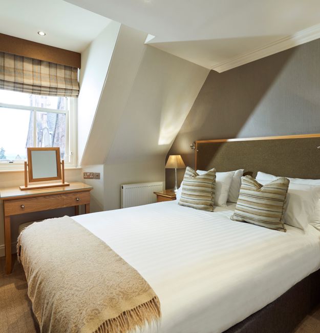 A room with double bed, vanity, bedside table, and dresser. The room has sloped ceilings.