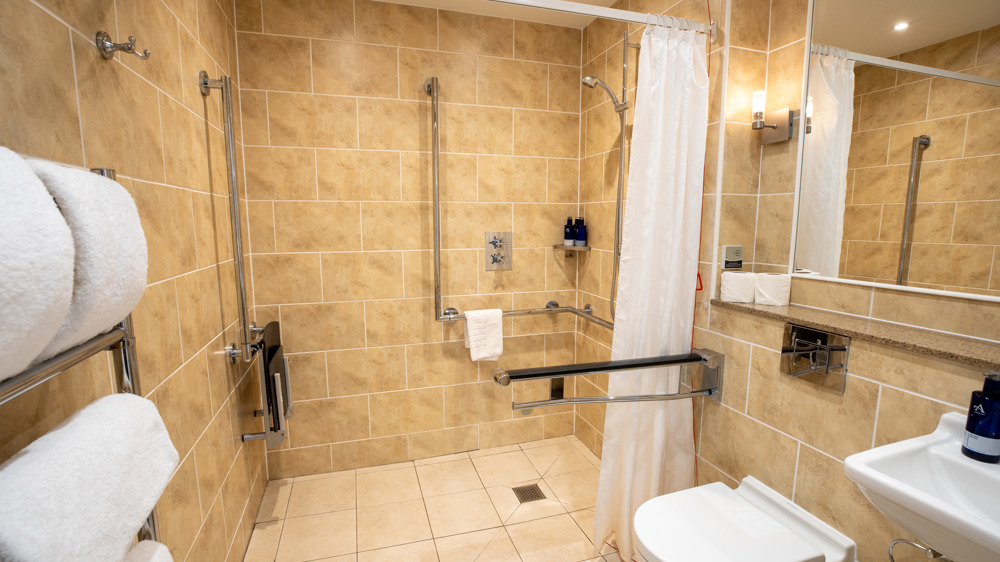 Accessible bathroom with roll-in shower with handles, toilet, sink, and towel rack.