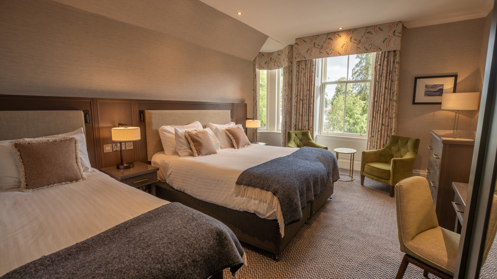 Room for 3 with a king-sized bed, a single bed, a vanity with chair, dresser, and two chairs next to a window.